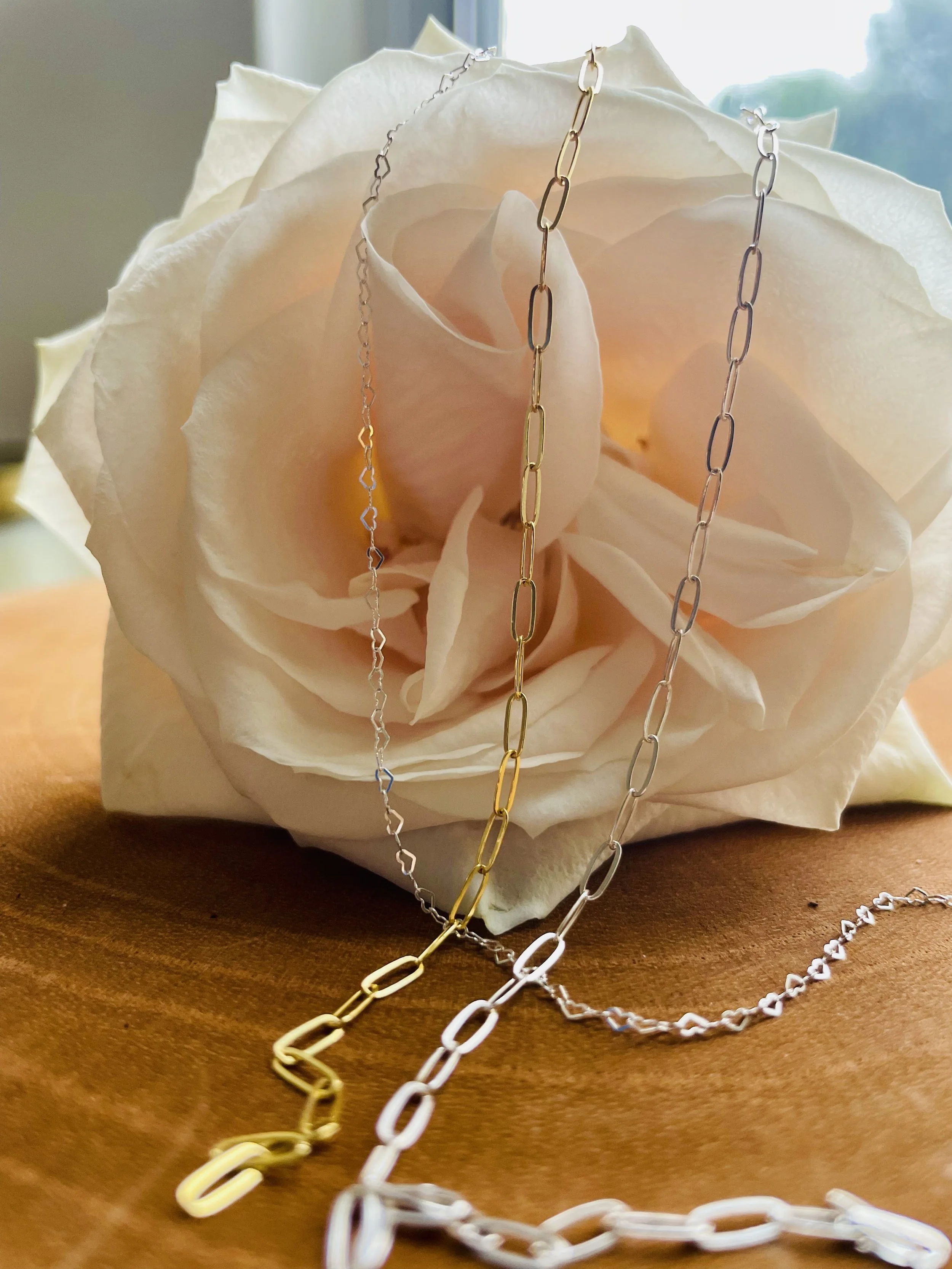 Three necklaces, silver, gold, and rose gold, draped over a white rose on a wooden surface.