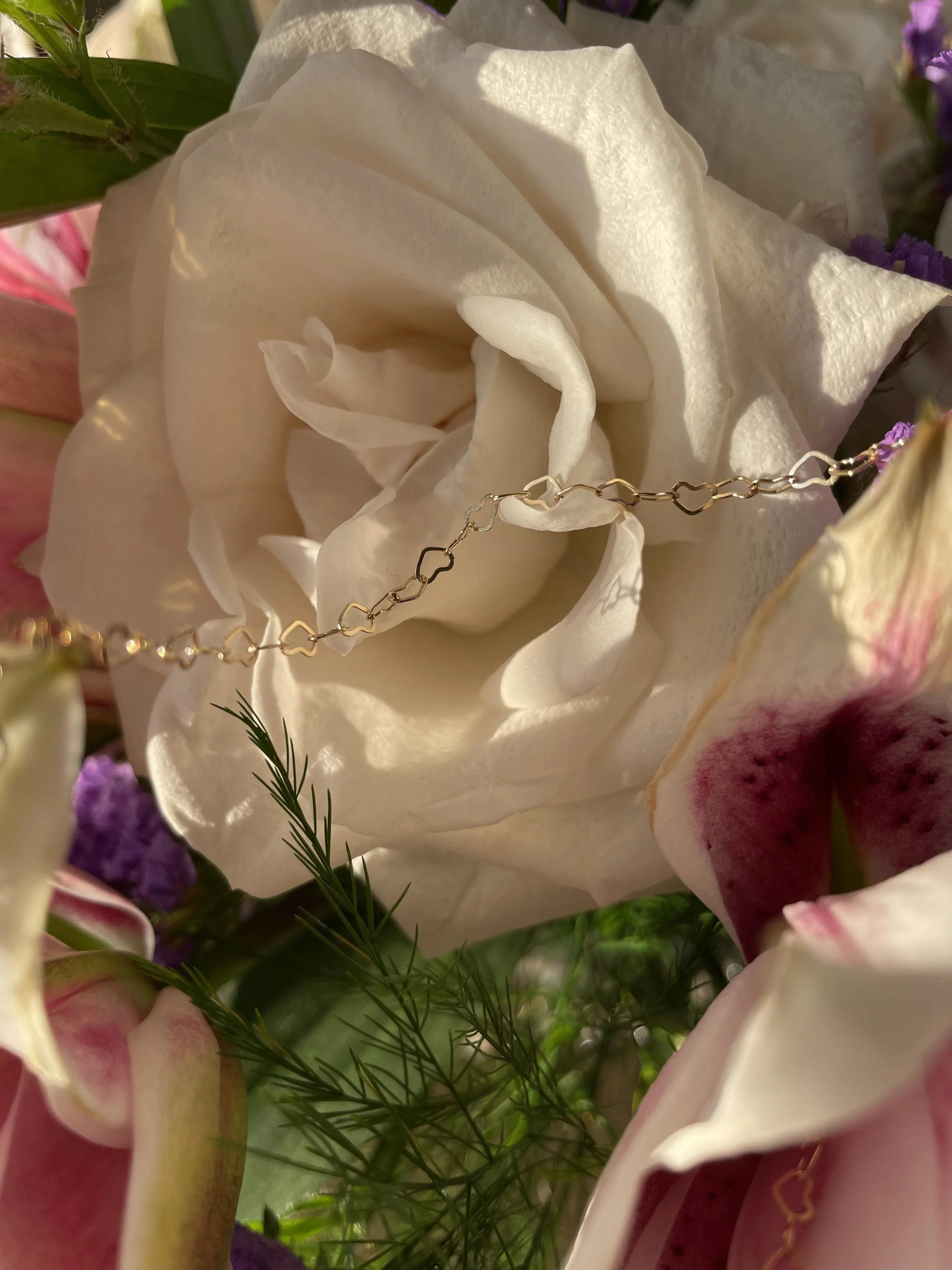 Close-up of a white rose surrounded by pink and purple flowers, with a delicate gold bracelet with small heart-shaped links draped across the bloom.