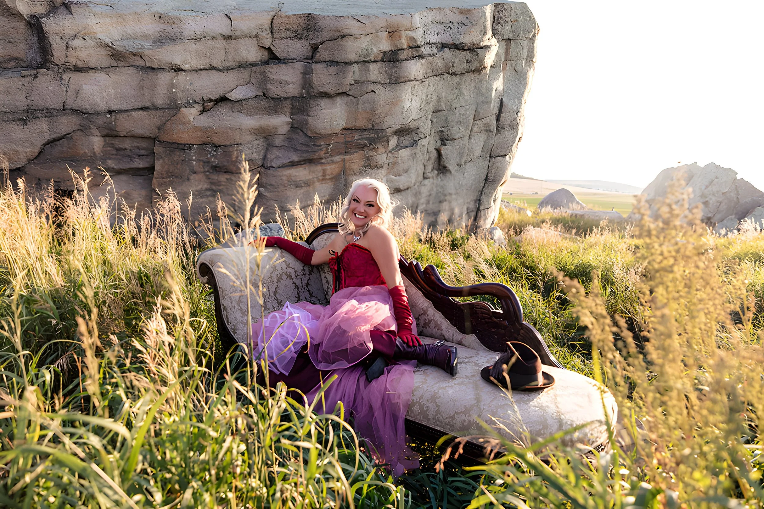 A woman in a red and purple dress sitting on a vintage sofa outdoors in a field of tall grass, with a large rock formation in the background and bright sunlight.