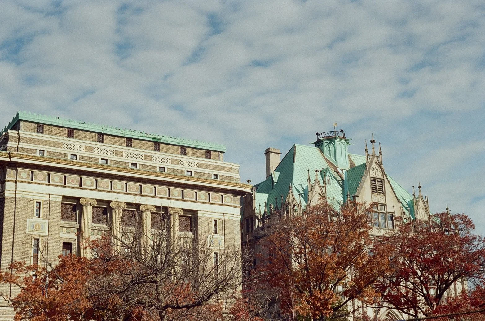 Historic buildings with green rooftops against a partly cloudy sky, surrounded by trees with autumn leaves.