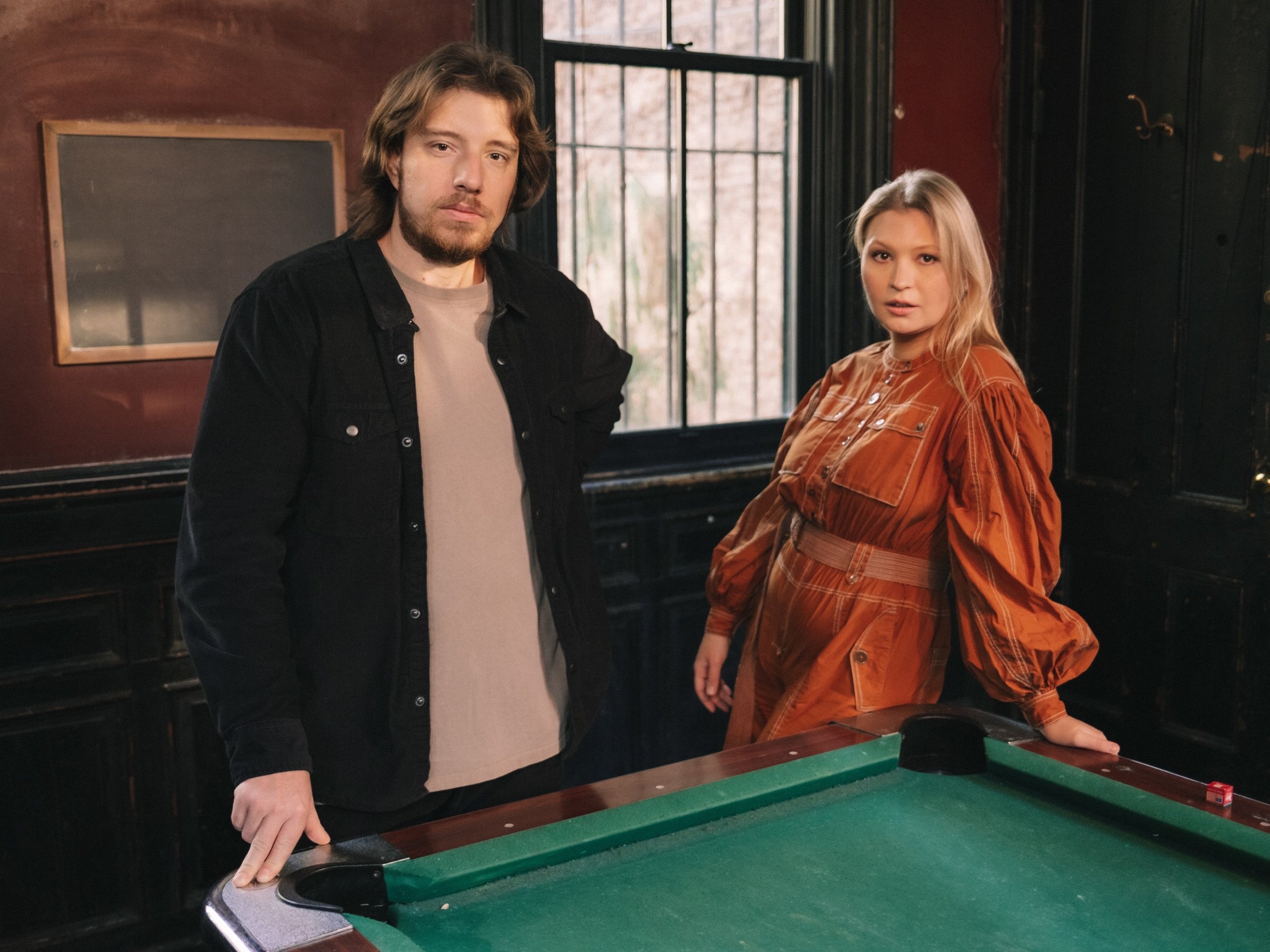 A man and a woman standing next to a pool table in a dimly lit room with wooden paneling and a large window in the background.