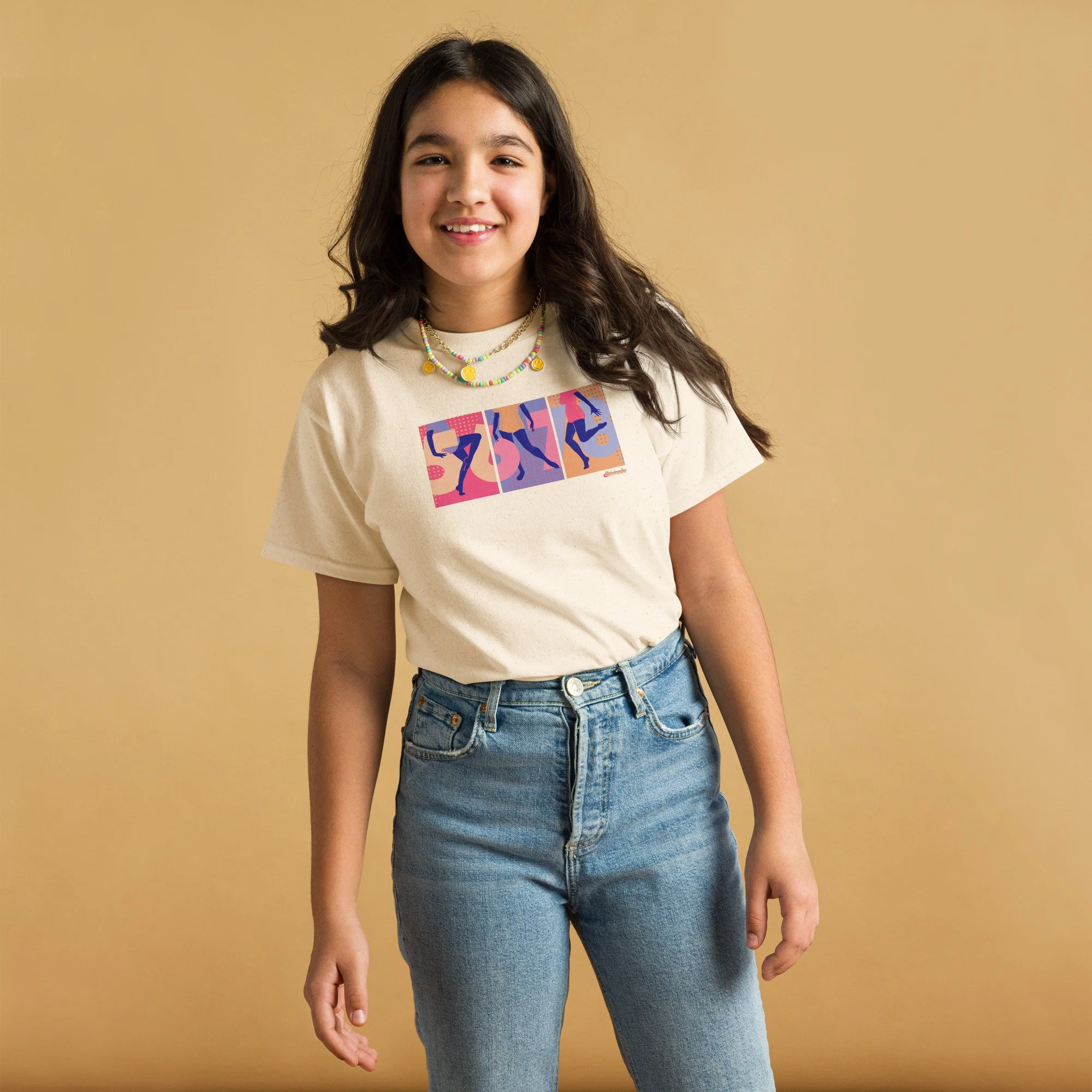 A teen girl with brown hair wearing a cream colored graphic tee with 5,6,7,8 and three dancers' legs spanning 3 panels, blue jeans.