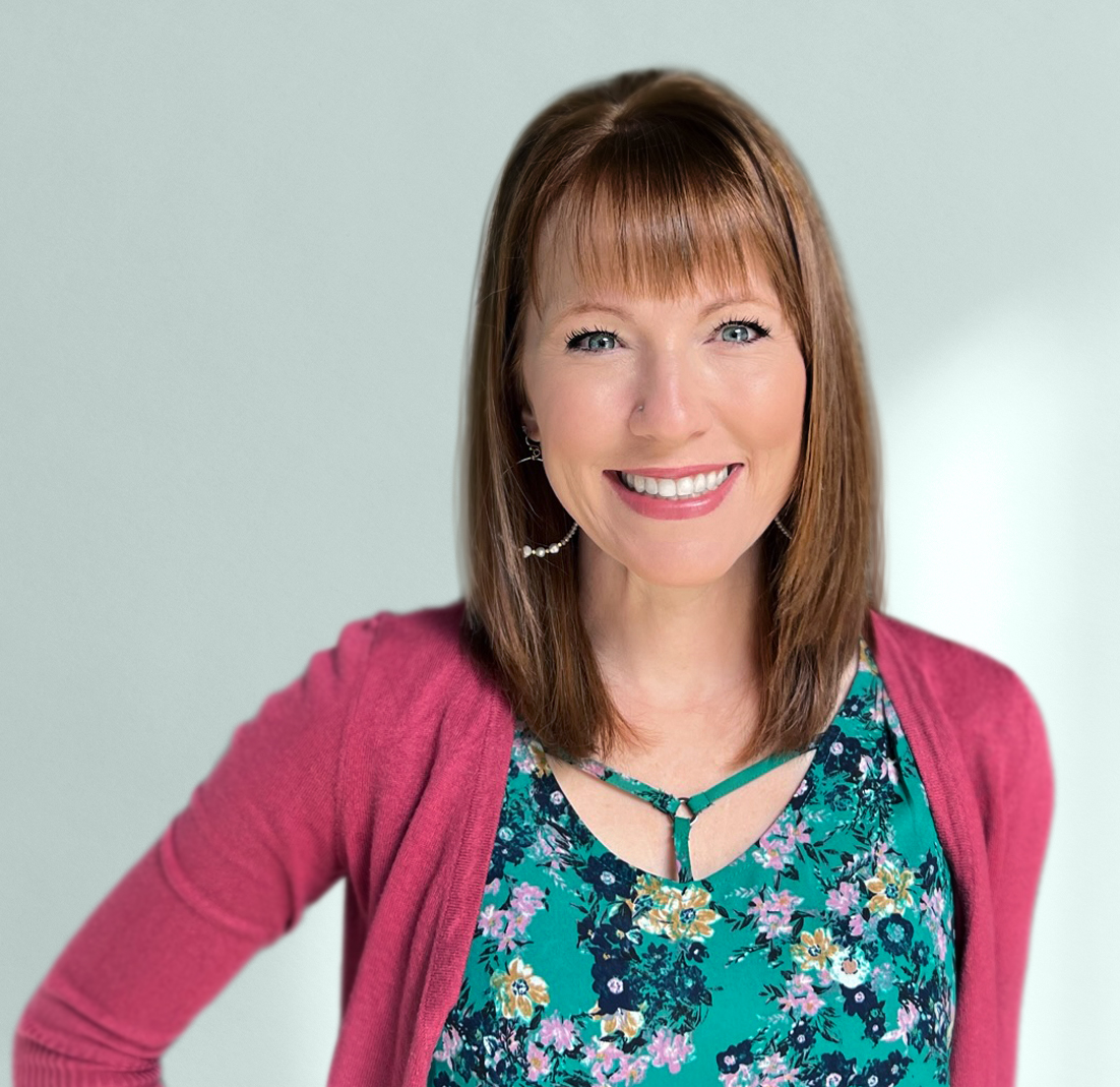 A woman with red hair, fair skin, and blue eyes, smiling outdoors in front of a barn-style door, wearing large white teardrop earrings and a blue sleeveless top with white embroidery.