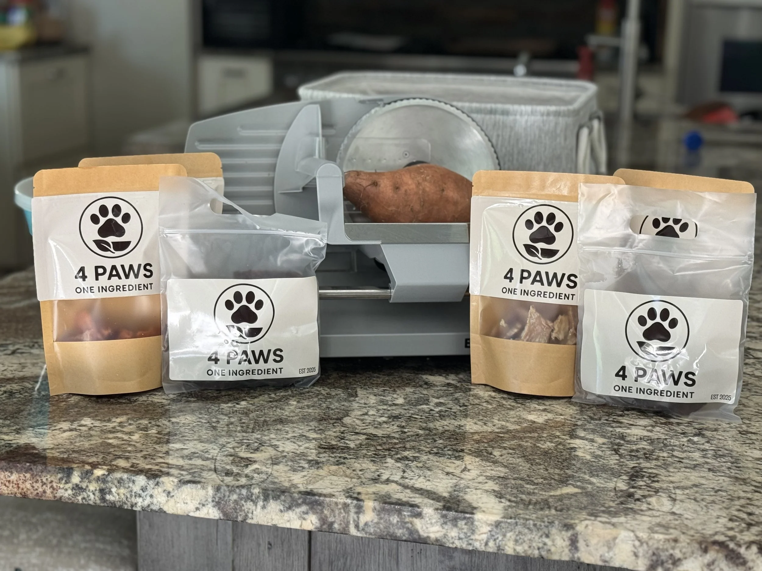Dog food packages labeled '4 PAWS, ONE INGREDIENT' on a kitchen countertop, with a raw sweet potato and a meat slicer in the background.