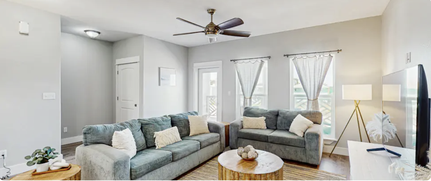 Living room with two gray sofas, a round wooden coffee table, a TV on a white stand, a standing lamp, and three windows with light curtains, ceiling fan, and light gray walls.