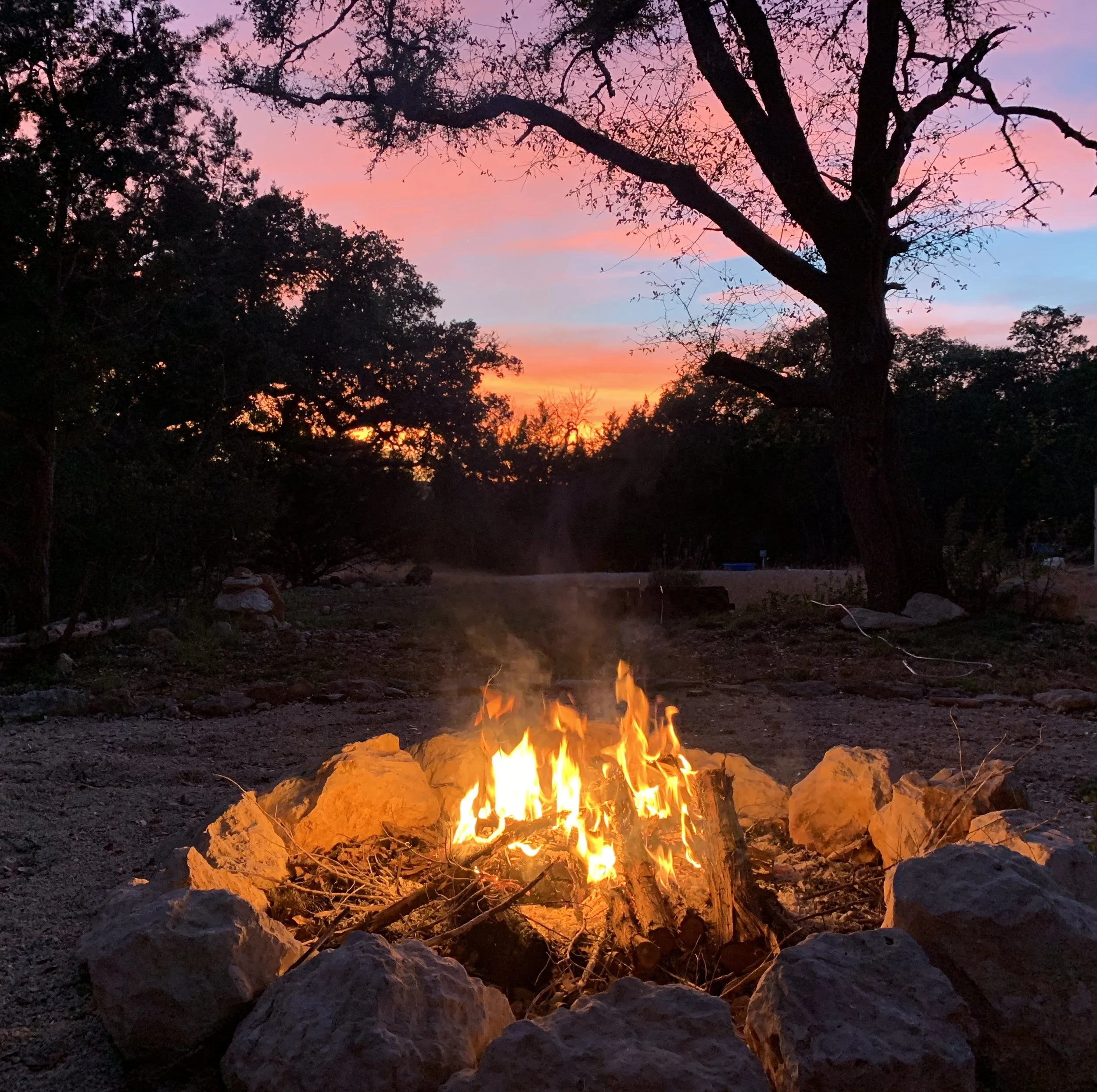 A campfire surrounded by rocks in an outdoor setting during sunset, with trees and a colorful sky in the background.