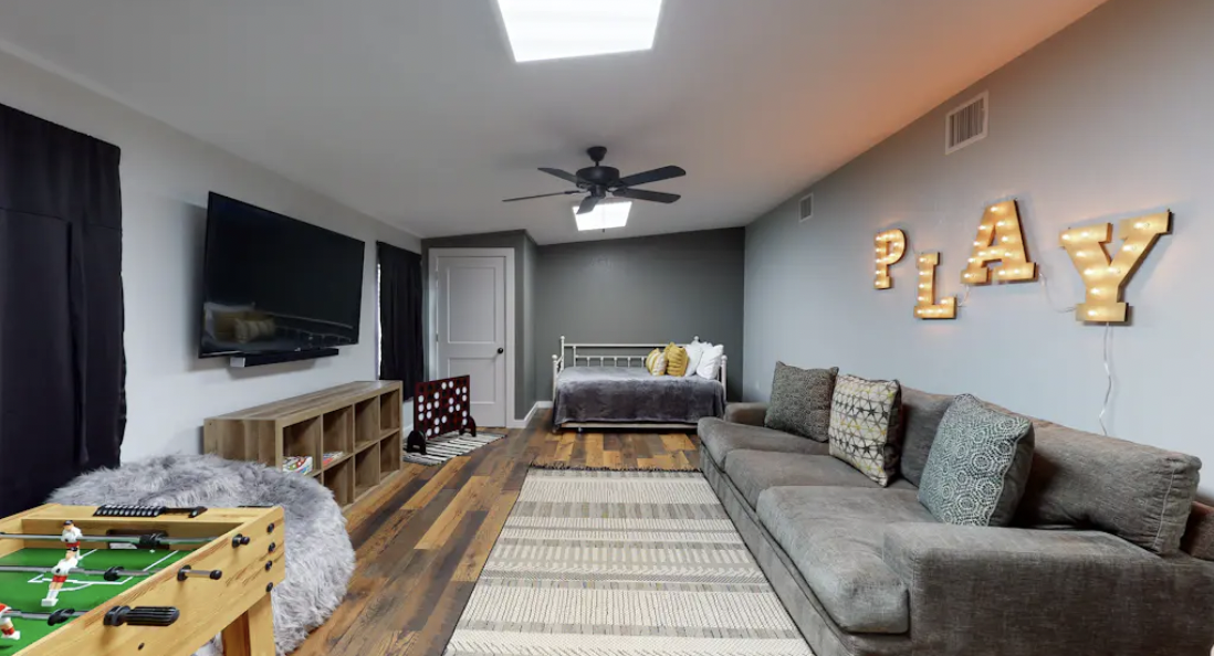 Living room with a gray couch, a television, a bed with pillows, and decorative letters spelling 'PLAY' on the wall.
