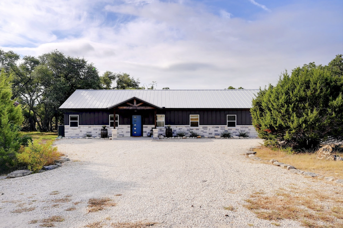 A long, single-story building with black siding, a metal roof, and a blue door, situated in a rural setting with trees and a gravel driveway.