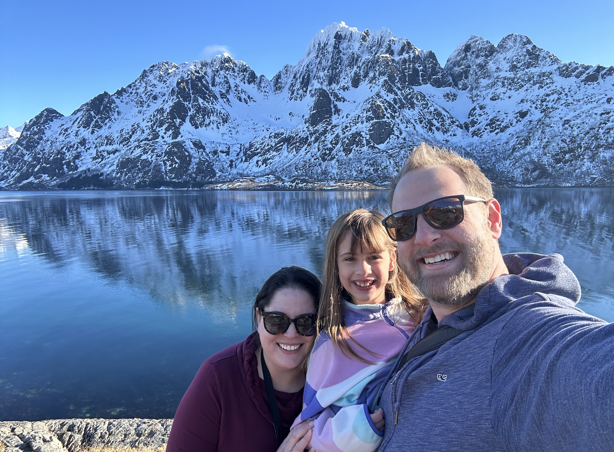 A smiling family of three taking a selfie by a lake with snow-capped mountains in the background on a clear day.
