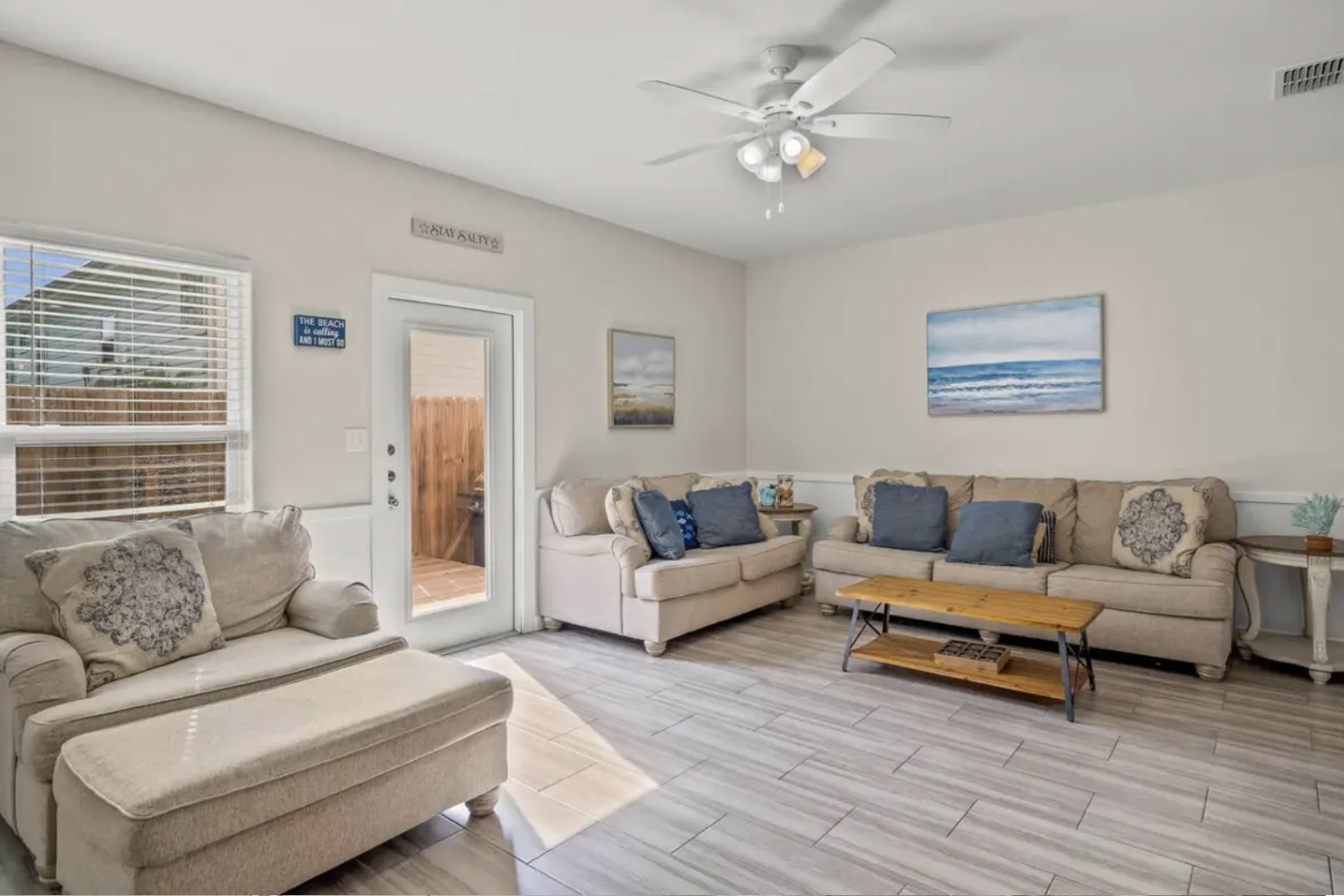Bright living room with beige sofas, blue cushions, a wooden coffee table, wall art of ocean scenes, window with blinds, door leading outside, ceiling fan with lights, and a sign that says 'Stay Salty'.
