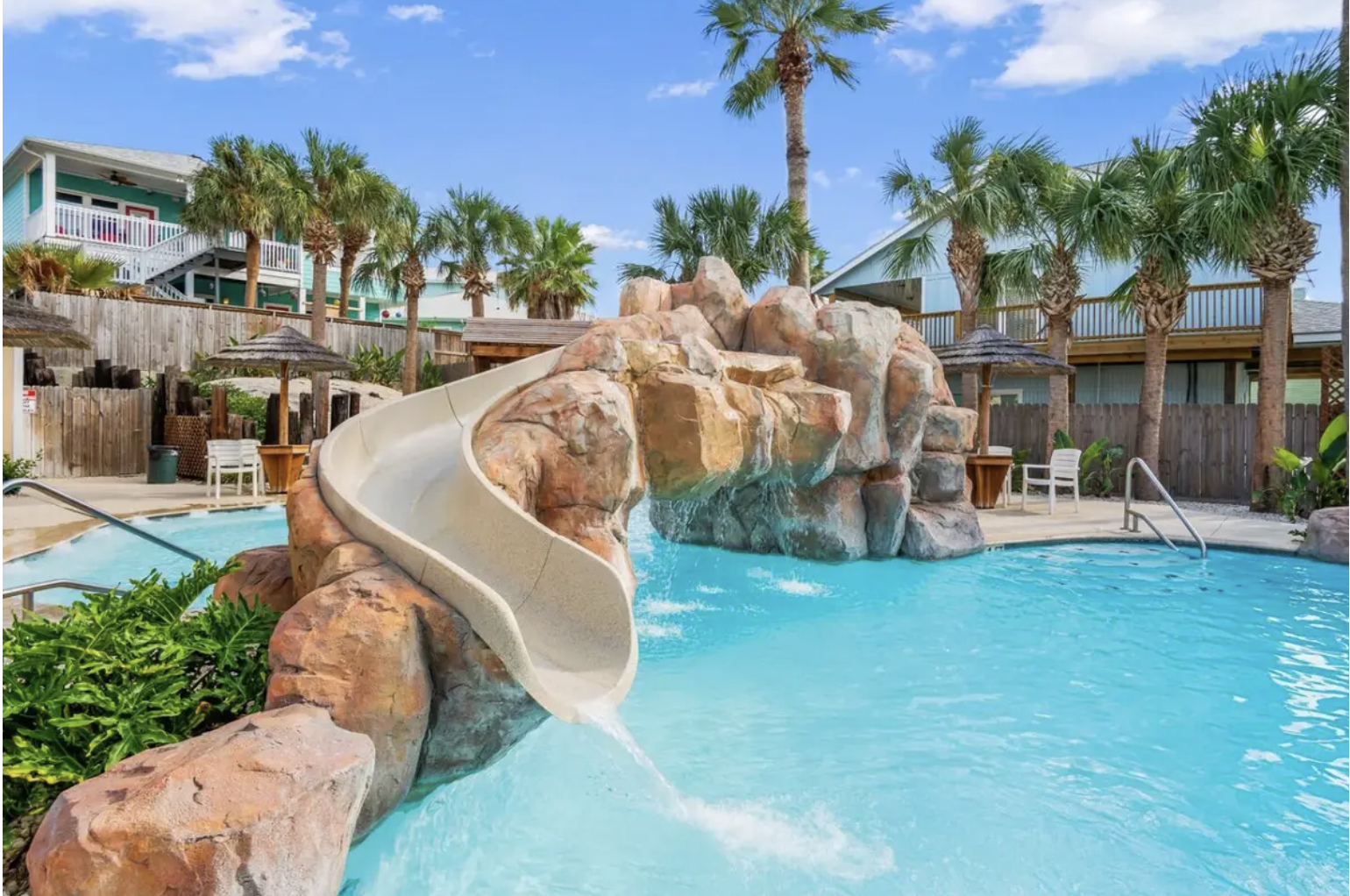 A swimming pool with a rock waterfall feature and a waterslide, surrounded by palm trees, lounge chairs, and wicker umbrellas, in a tropical resort setting.