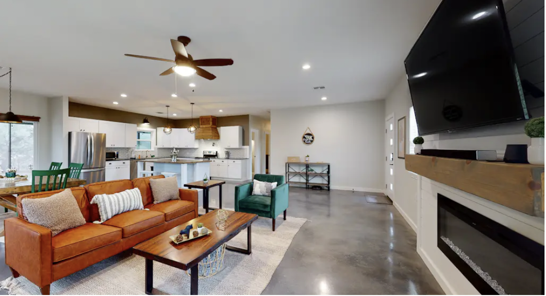 Living room with orange sofa, green armchair, wooden coffee table, electric fireplace, mounted TV, and open kitchen in the background.