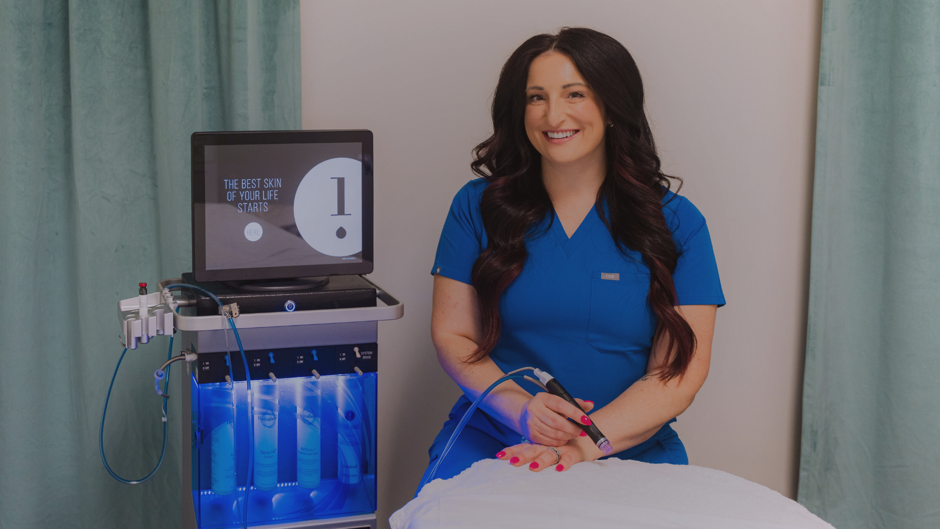A smiling woman in blue scrubs sitting in a medical setting next to a Hydrafacial machine, holding a hand-held device, with a monitor displaying skincare message.