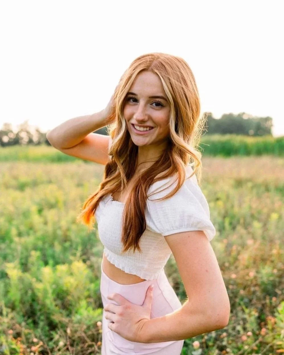 A front desk/receptionist at skin lab aesthetics & wellness with long, wavy red hair smiling and posing outdoors in a field during daytime.