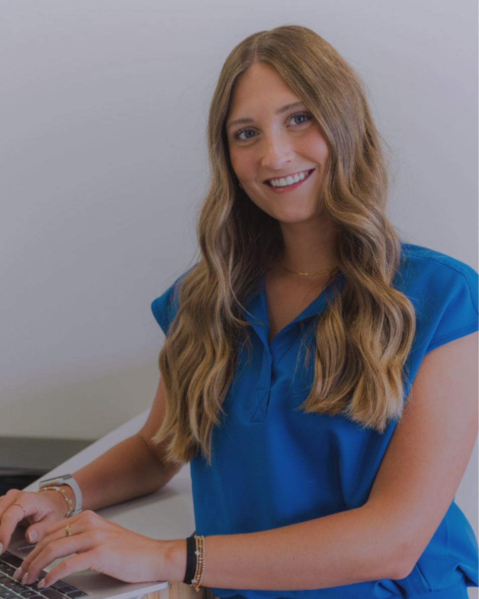 The practice manager at skin lab aesthetics & wellness with long wavy hair wearing a blue shirt, sitting at a desk, using a laptop, smiling at the camera.