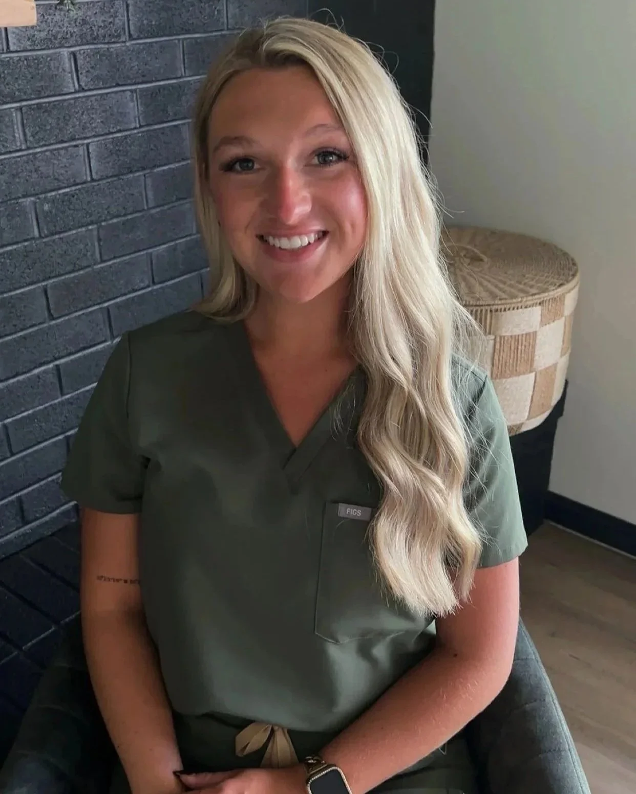 A certified nurse injector at skin lab aesthetics & wellness with long, wavy blonde hair smiling, wearing green medical scrubs sitting indoors against dark brick wall and a piece of wicker furniture.