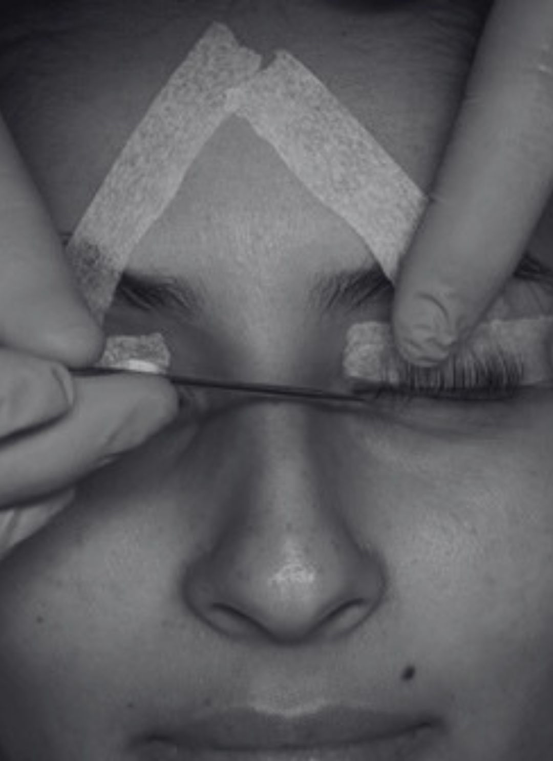 Close-up of a person's face with eyes closed, receiving a lash lift and tint, with a person's hands gently holding the skin and using a tool on the lashes.