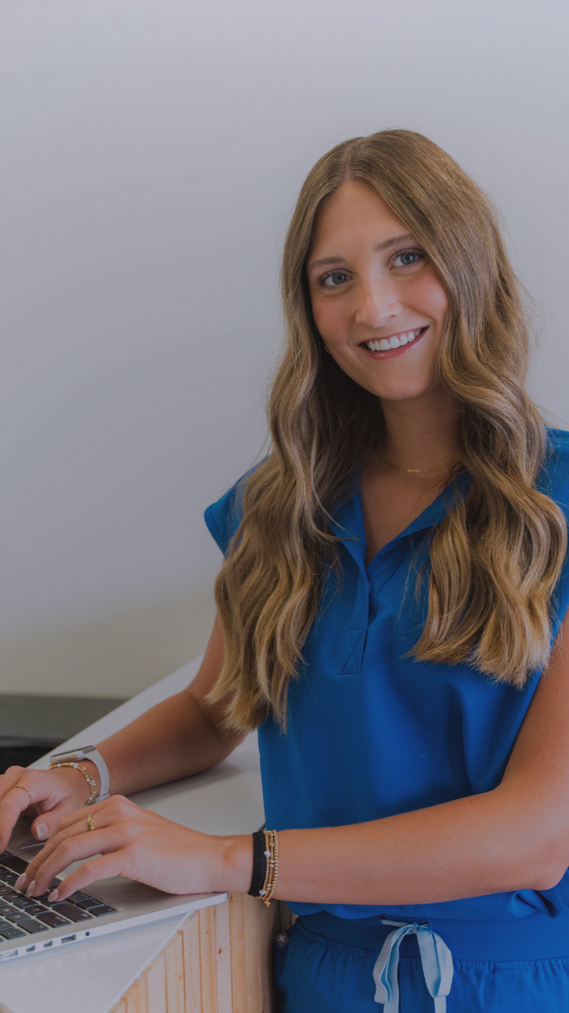 A young woman with long wavy brown hair, smiling, wearing a blue collared shirt and gold jewelry, sitting at a desk and working on a laptop.
