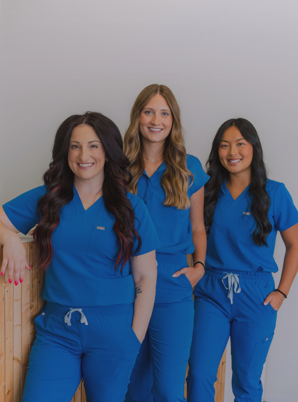 Three women in blue scrubs standing together indoors, smiling at the camera.