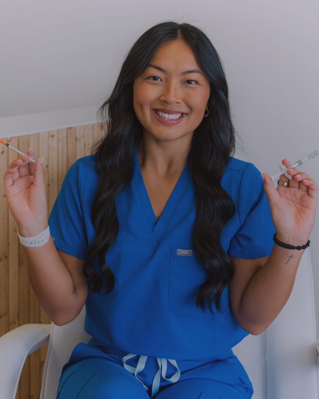 A nurse in blue scrubs holding a syringe, smiling, inside a medical facility.