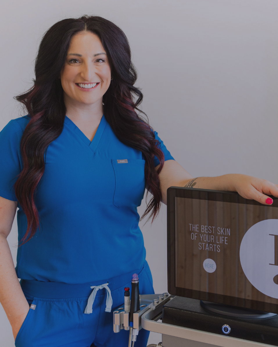 A smiling woman in blue medical scrubs standing next to a monitor displaying skincare promotion text. She has long dark hair with red highlights and is holding a medical device with pens in the pocket of her scrubs.