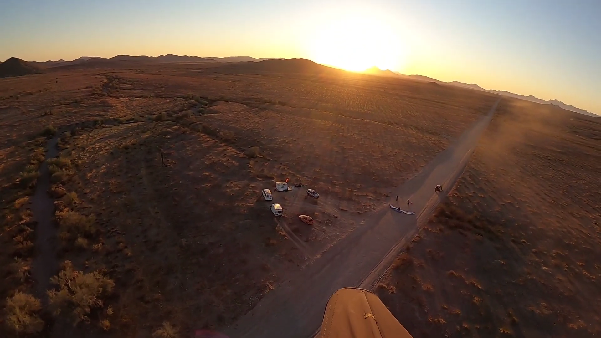 Aerial view of a desert landscape at sunset with a dirt road, parked vehicles, and scattered trees from the cockpit of a paraglider that is descending