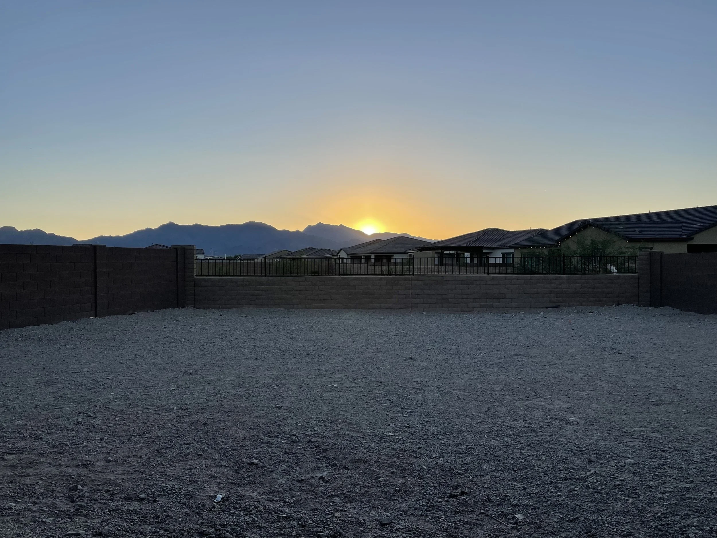Backyard with unimproved gravel ground, brick wall view fence, neighboring houses, and mountains with a sunrise/sunset in the sky.