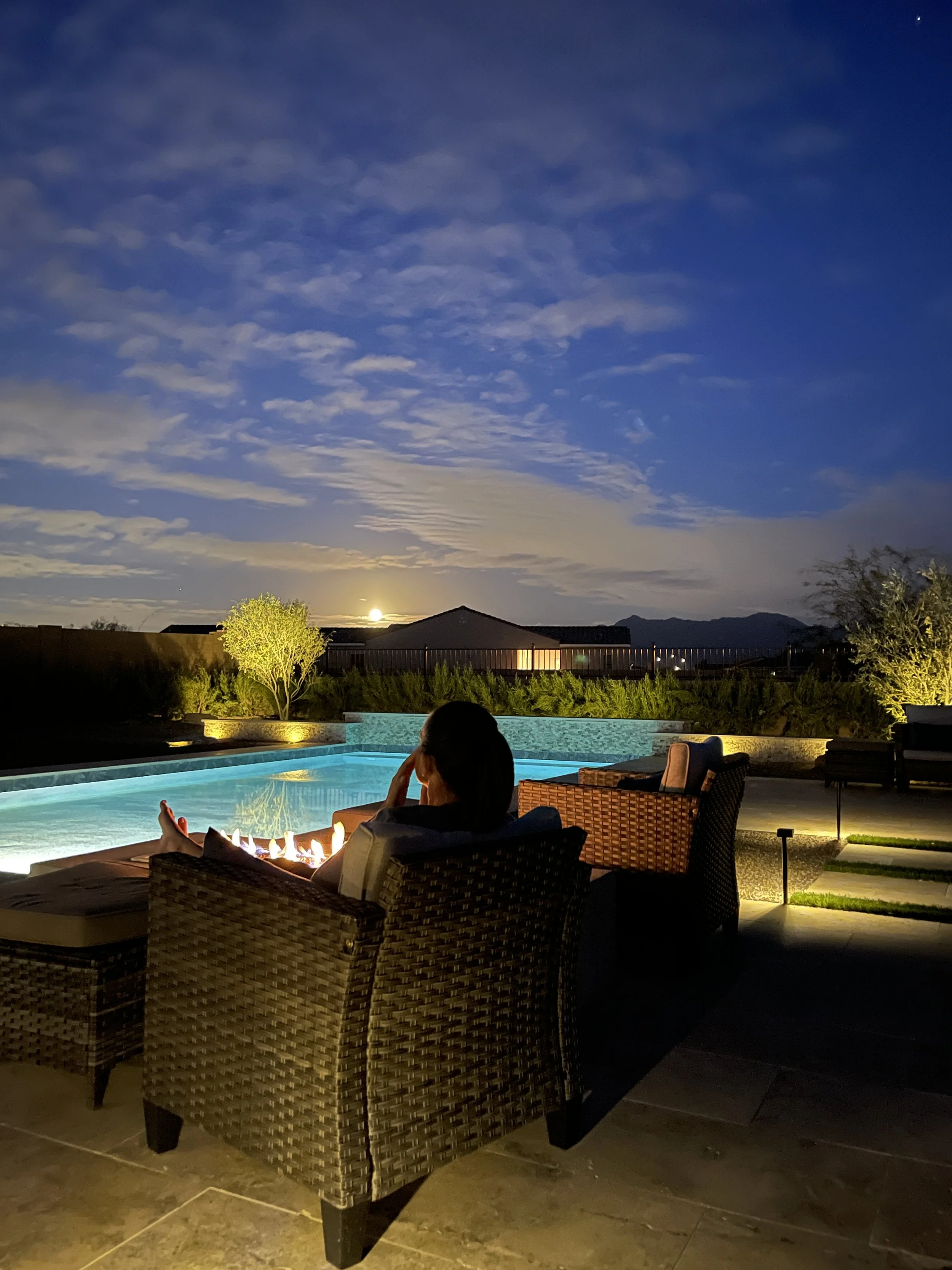 A woman relaxing by a backyard swimming pool at night, with a fire pit, illuminated chairs, and a view of the moon in the sky.
