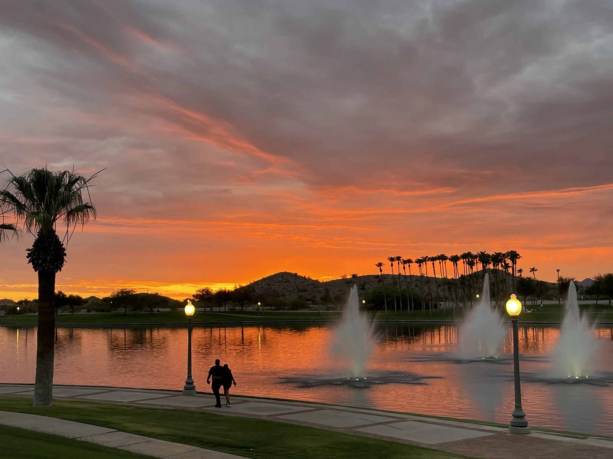 A sunset over a park with palm trees, a lake with fountains, and a couple walking near the water, with street lamps lit.