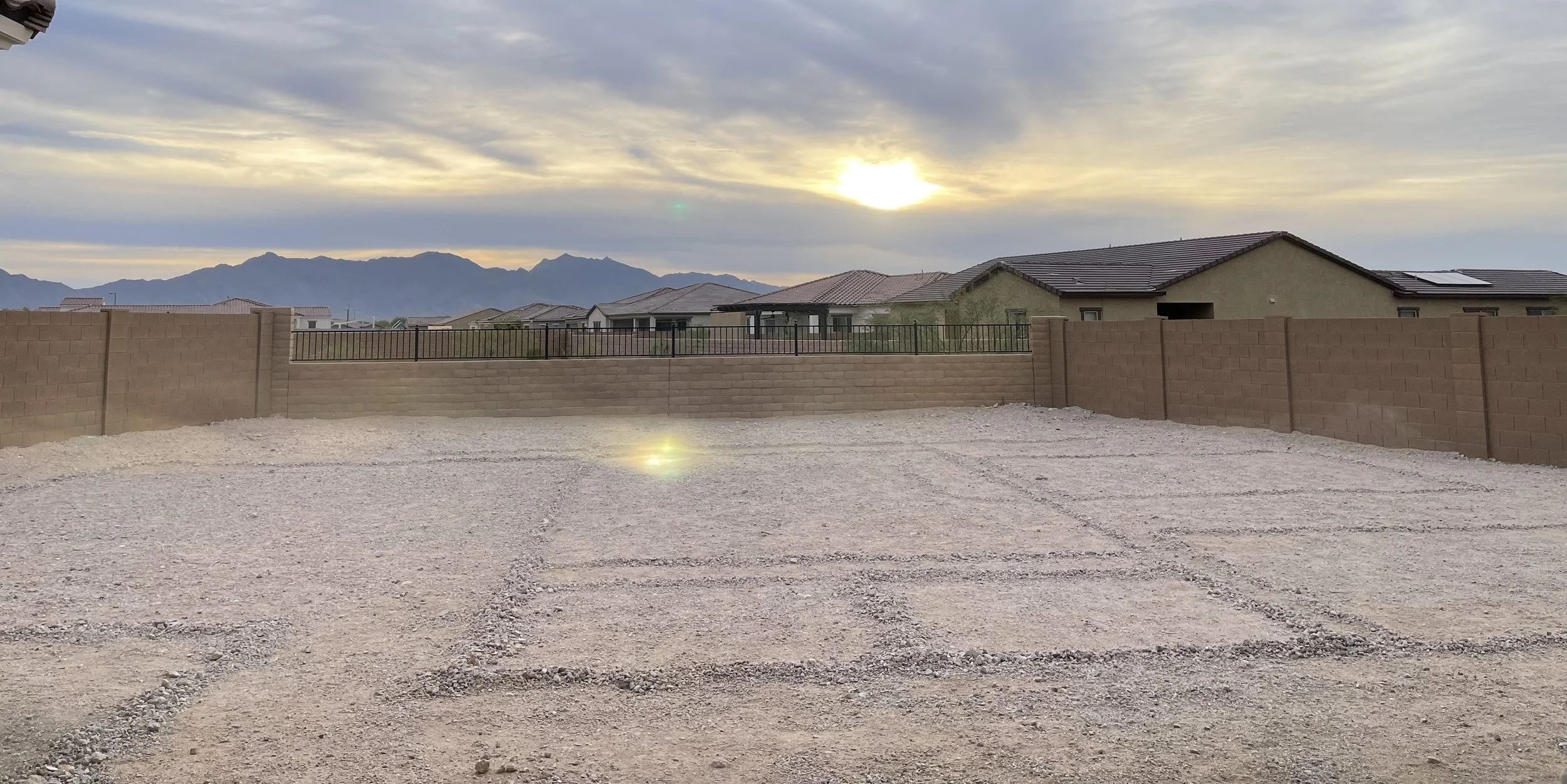 Empty gravel parking backyard lot with a brick wall and neighborhood houses, mountains in the background, and a partly cloudy sky with the sun near the horizon.
