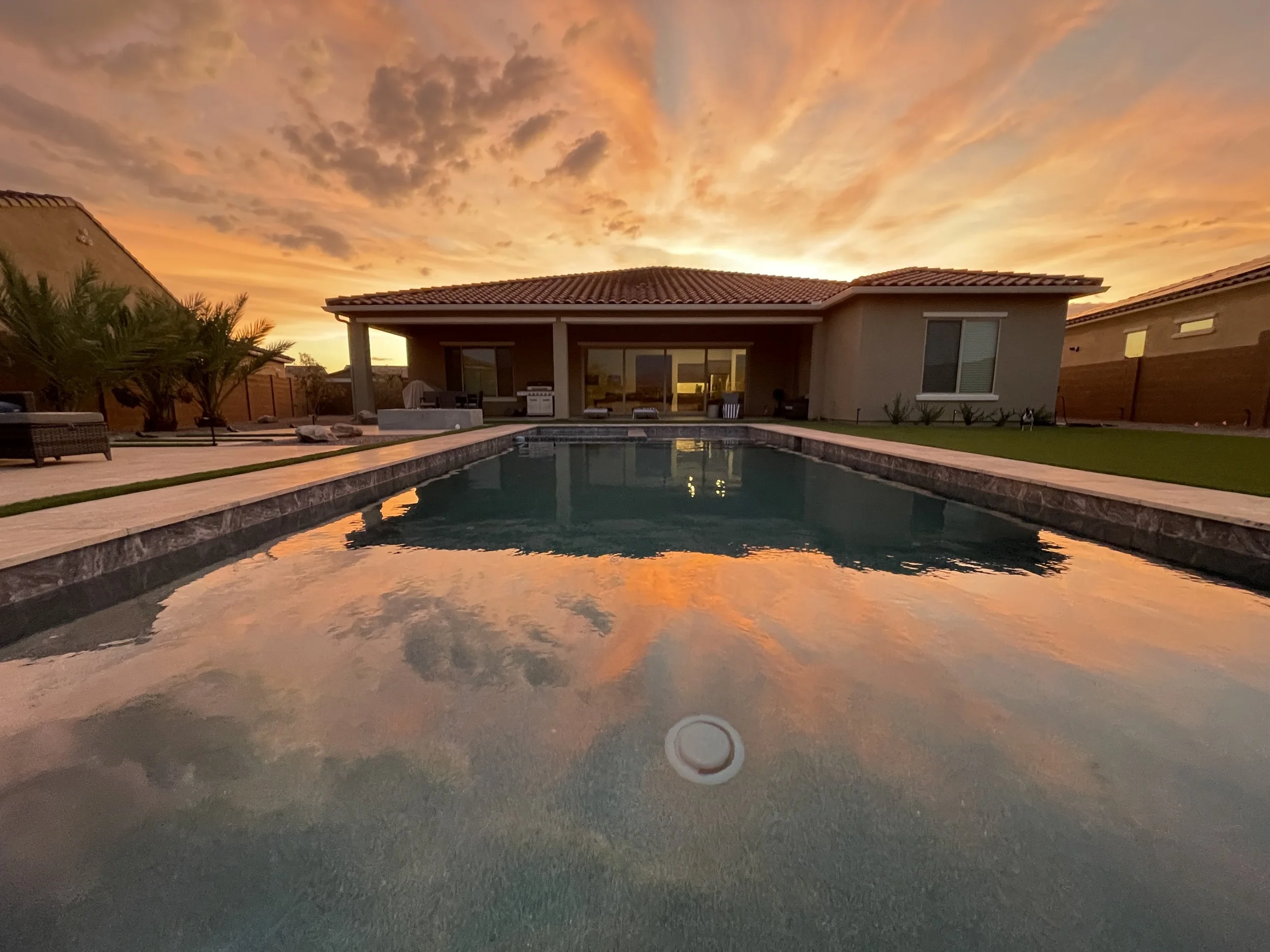 A backyard with a swimming pool reflecting the colorful sunset sky, a house with a covered patio, outdoor furniture, and some landscaped greenery.
