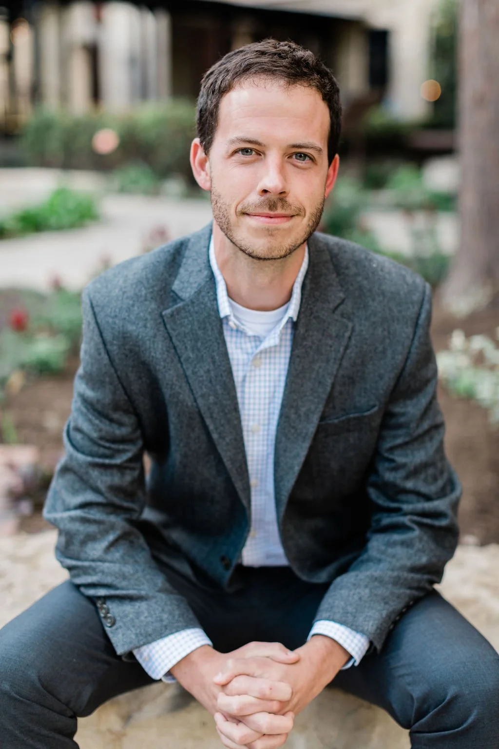 A man in a gray blazer and checkered shirt sitting outdoors in a garden area with blurred plants and structures in the background.