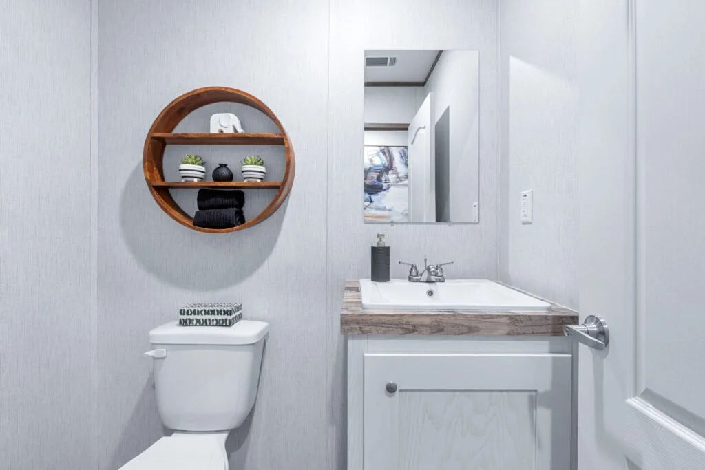 A small bathroom with light-colored walls, a round wooden shelf with plants and decorative items, a mirror above a small sink with a wooden countertop, a soap dispenser, a white toilet with a patterned lid, and a door on the right side.