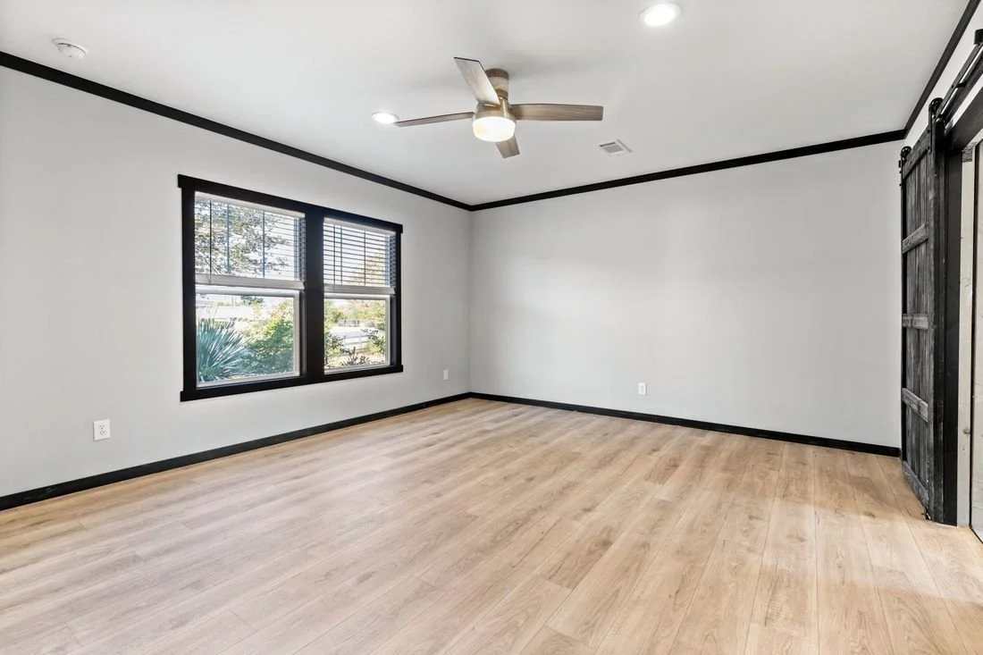 Empty room with two windows, light wood flooring, white walls with black trim, a ceiling fan, and a rustic sliding barn door.