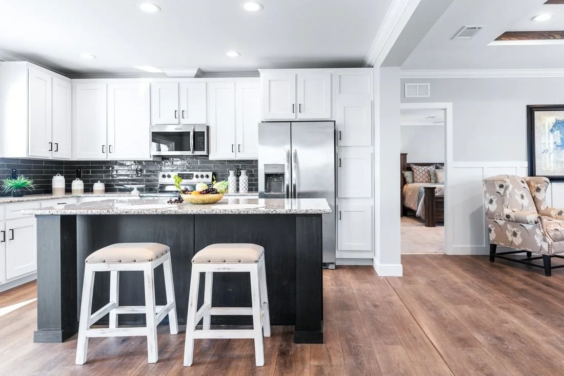 Modern kitchen with white cabinets, stainless steel refrigerator, black subway tile backsplash, granite countertop, and an island with two white stools. Adjacent living space features a patterned armchair and a bedroom visible through an open doorway