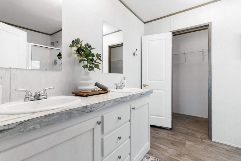 A bathroom with a marble countertop, a double sink, a large mirror, a white door, and an open closet with a hanging rod. There is a plant on the counter and a towel.