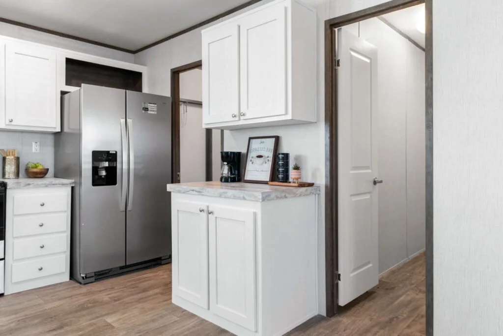 A kitchen with white cabinets, a stainless steel refrigerator, and a small counter with a coffee maker, framed print, and decor. Wooden flooring and two doorways, one leading to a pantry or closet and the other to a hallway.