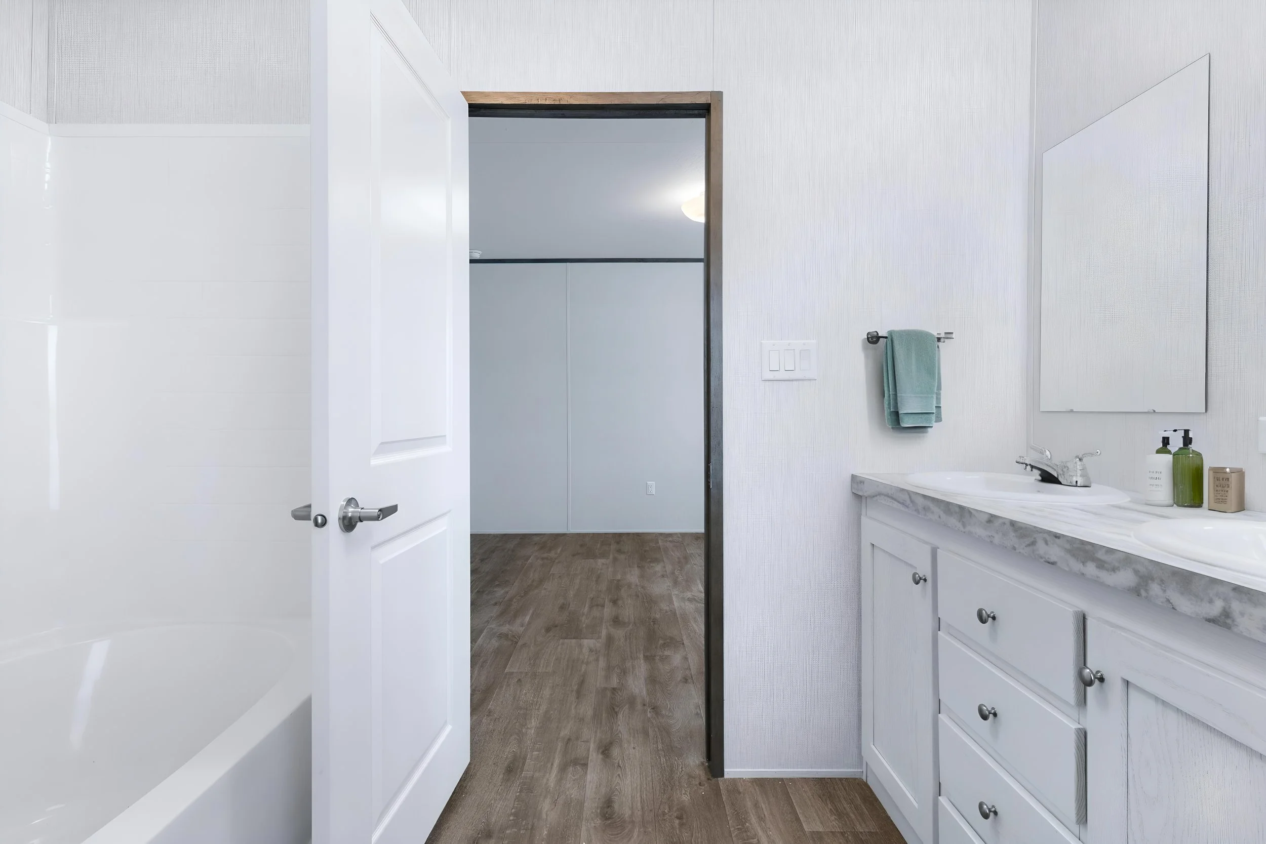 A modern bathroom with white walls, a marble countertop, and a white sink. There are bottles on the countertop, a towel hanging on a bar, and a mirror on the wall. An open door reveals a closet or another room, and the floor is made of wood-like mate