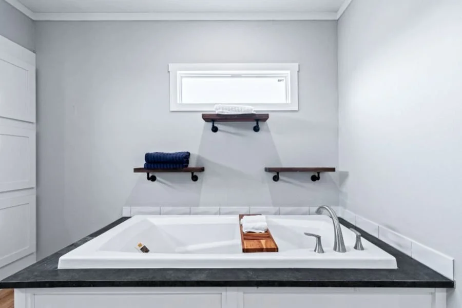 White bathtub with black countertop, three wooden shelves above, a small window, and a towel tray with towels inside the tub.