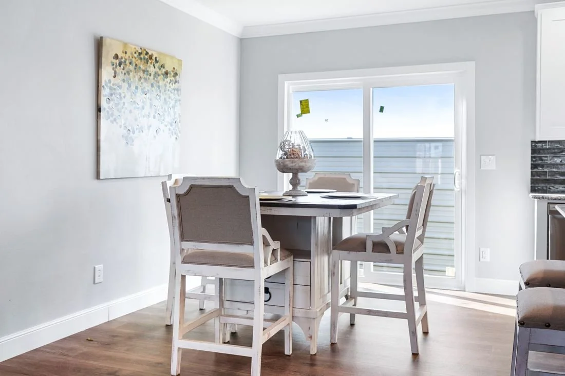 Dining area with a white wooden table, four beige chairs, a glass jar decoration, and a large window with a door leading outside