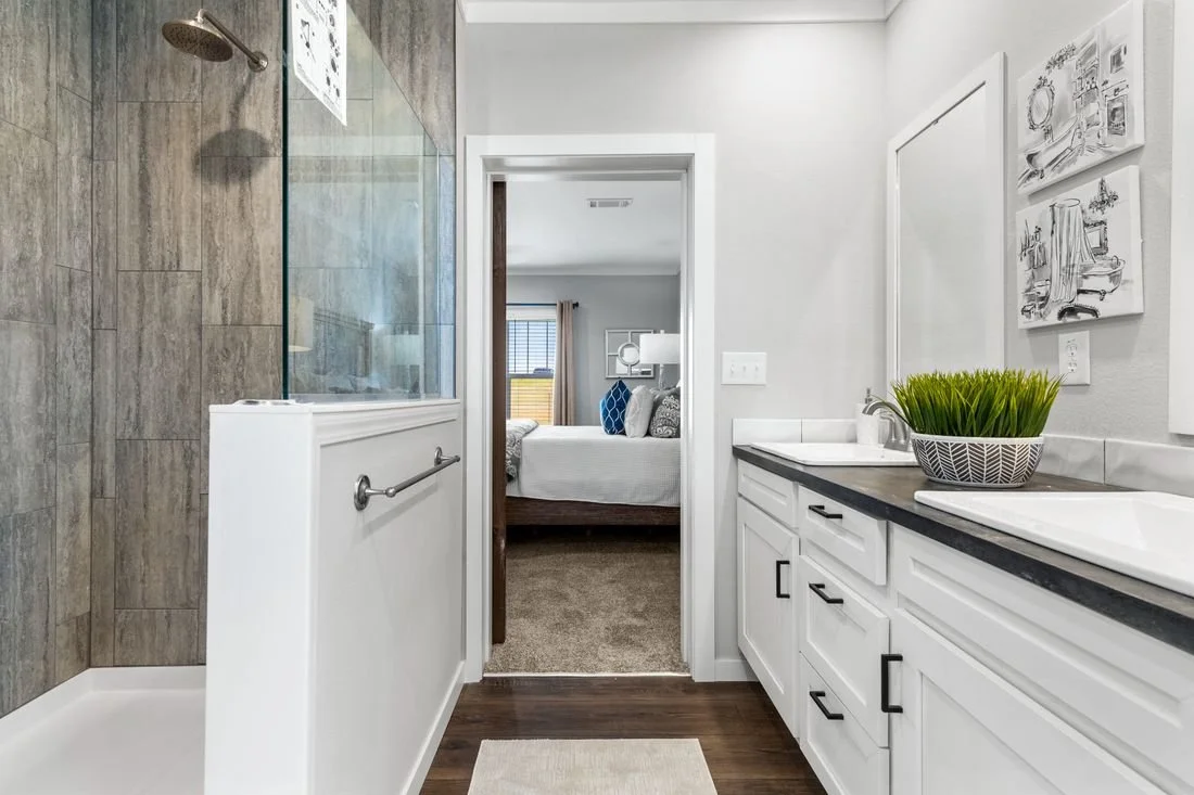 View of a bathroom with a shower on the left, white double-sink vanity with black countertop on the right, and a doorway in the center leading to a bedroom with windows and a bed.