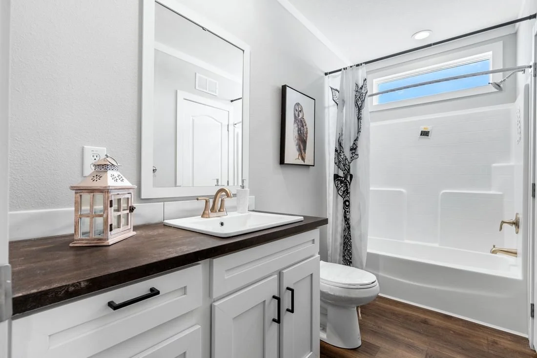A modern bathroom with a wooden countertop, white cabinetry, a white sink with gold fixtures, a mirror, a framed owl picture, a lantern decoration, a bathtub with a curtain, and a small window at the top.
