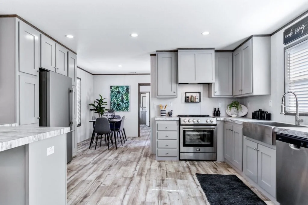 Modern kitchen with gray cabinets, stainless steel appliances, farmhouse sink, and light wood flooring. There is a window with blinds, a wall chalkboard, and a dining area with a round table and chairs in the background.