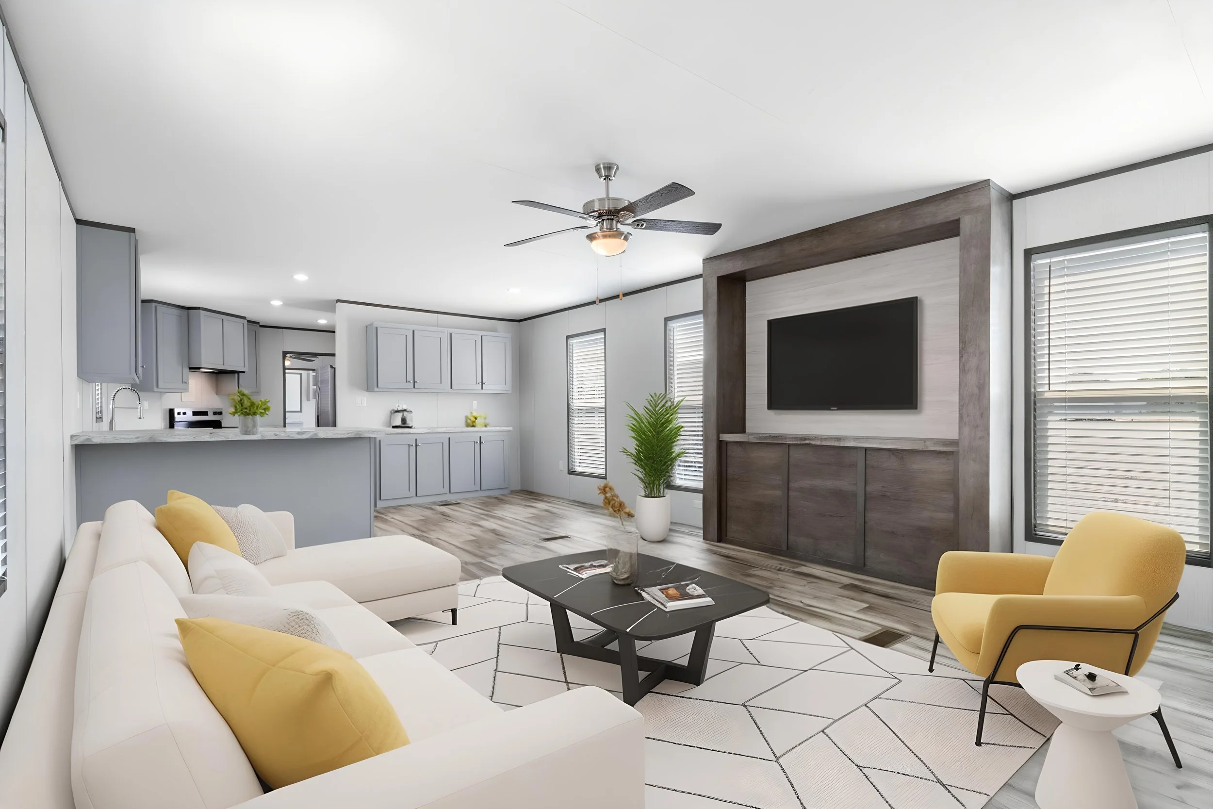 Open-concept living room and kitchen with light-colored furniture, yellow accent pillows, a black coffee table, a mounted flat-screen TV, ceiling fan, and large windows with blinds.