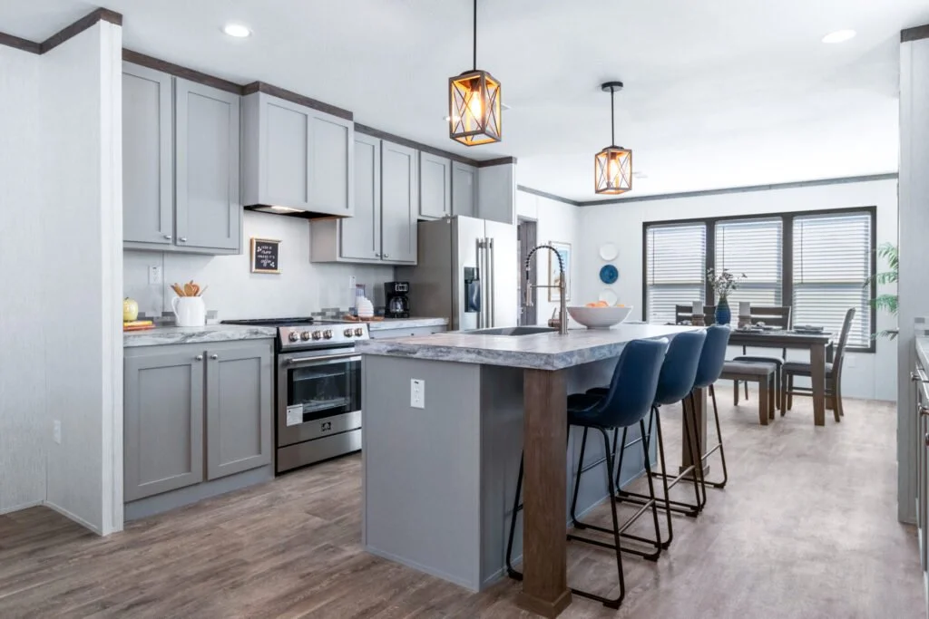 Modern kitchen with light gray cabinets, stainless steel stove, refrigerator, and black coffee maker. Marble countertop island with four navy blue bar stools. Dining area with large windows, wooden table, and chairs, decorated with vases and artwork.
