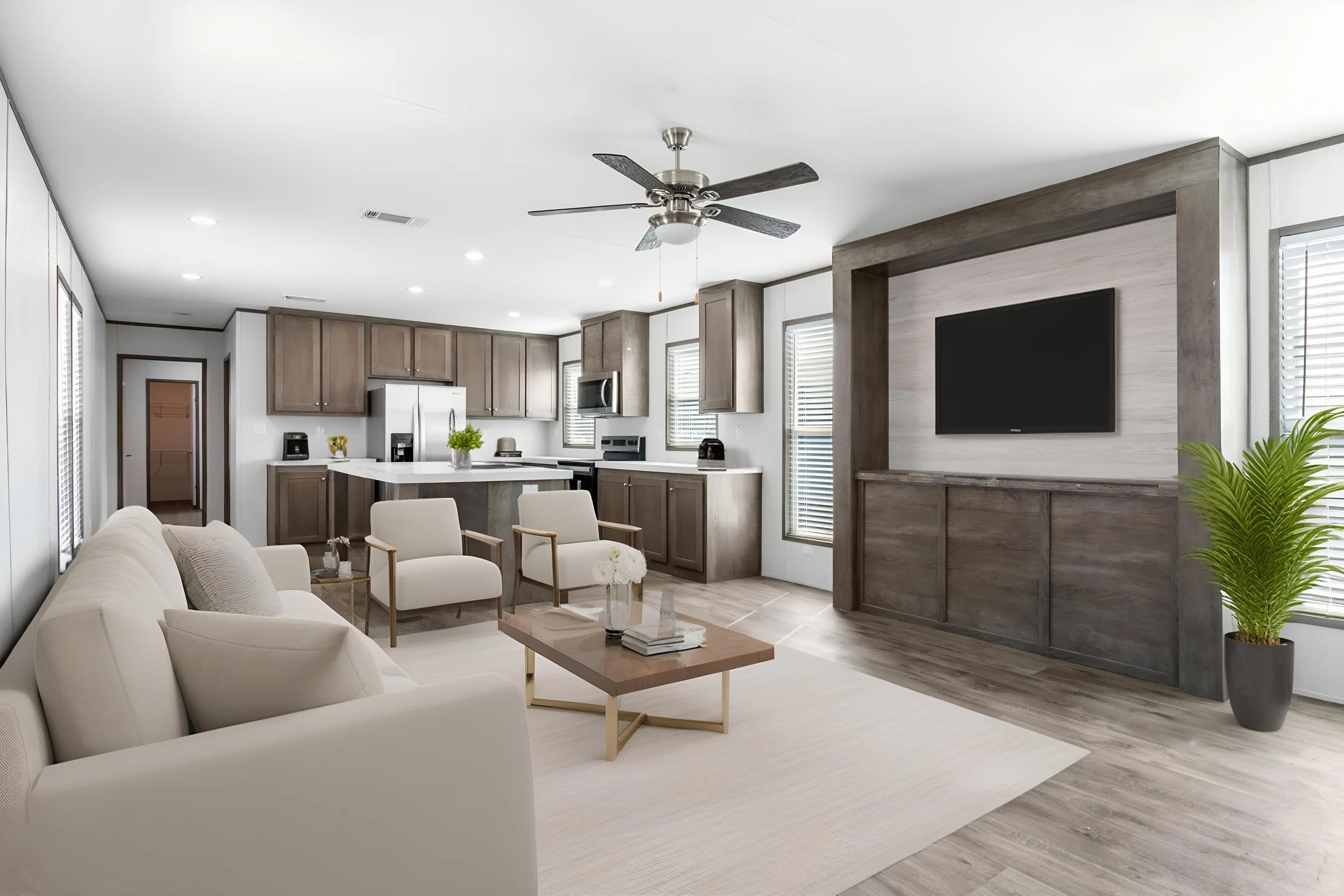 Open-concept living room and kitchen with white sofa, beige armchair, wooden coffee table, kitchen cabinets, white countertop, stainless steel appliances, ceiling fan, flat-screen TV on a wooden accent wall, multiple windows with blinds, and a potted plant.