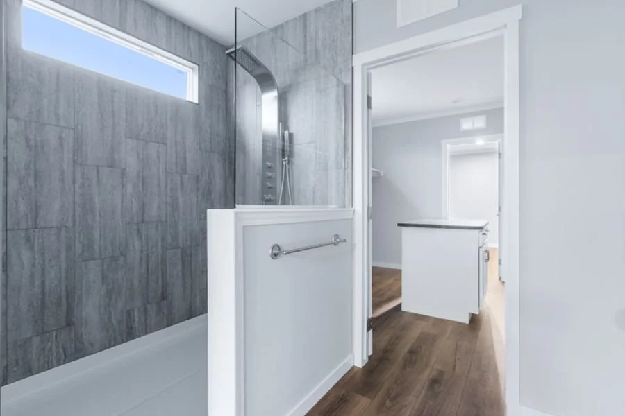 Bathroom with a walk-in shower, gray tiled walls, a small window, a towel bar, and white trim. A doorway leads to another room with hardwood floors.
