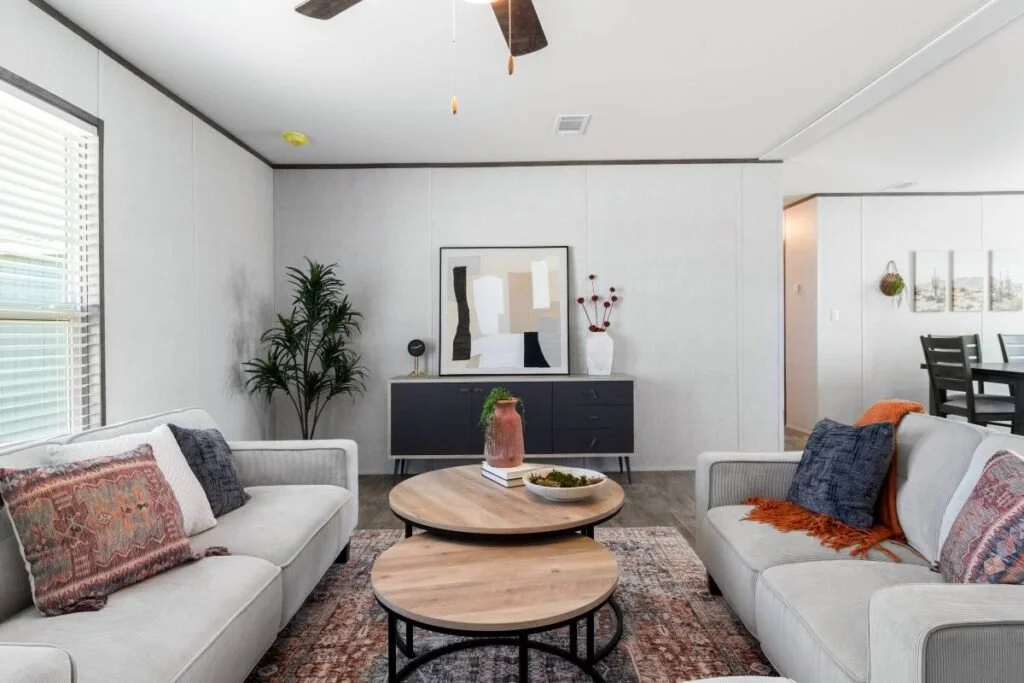 Living room with two white sofas, patterned pillows, round wooden coffee tables, a large abstract painting, a tall plant, and a sideboard with vases and decorative items.