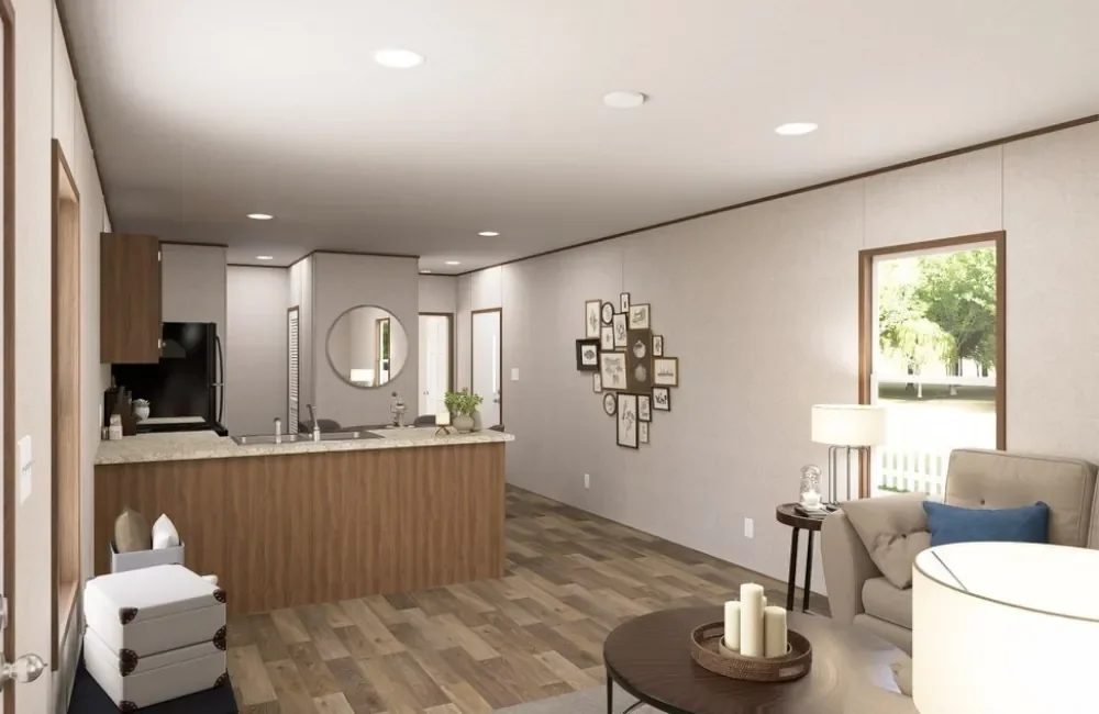 Open-concept living room and kitchen with a neutral color scheme, wood flooring, and minimal decor. The living area has a beige armchair with a blue pillow, a side table with a lamp and decorative items, and a round coffee table with candles. The kit