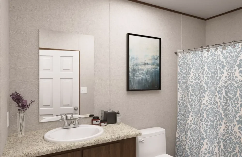 Bathroom with beige walls, a white vanity with a granite countertop, a sink, a mirror, a toilet, a curtain with a gray and white floral pattern, a framed painting, and decorative items on the countertop.